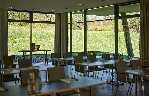 Mehrere Reihen von Tagungstischen im Saal mit großen Fenstern und Blick auf den Garten