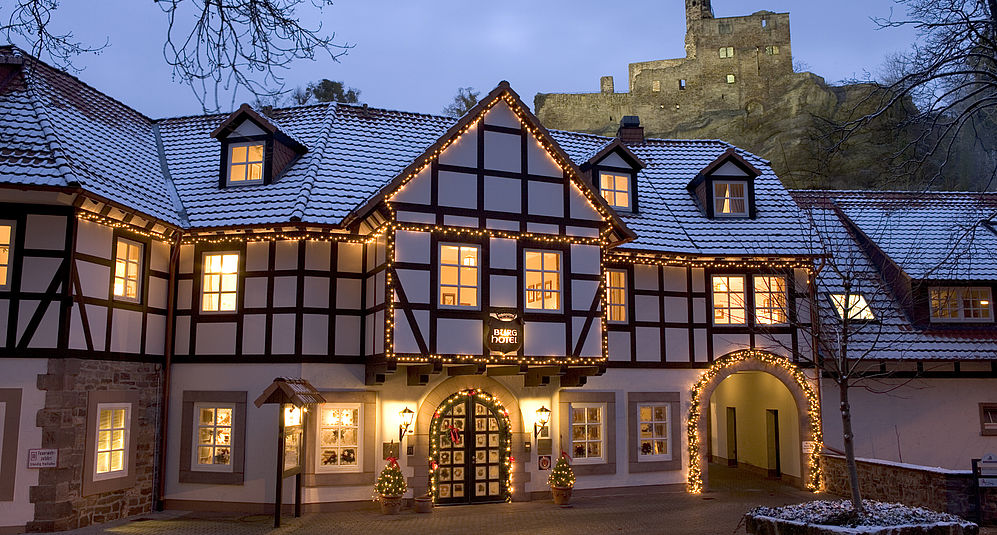 Beleuchtete Fachwerkfassade des Hardenberg BurgHotels im Winter, dahinter eine beleuchtete Burgruine bei Abenddämmerung.
