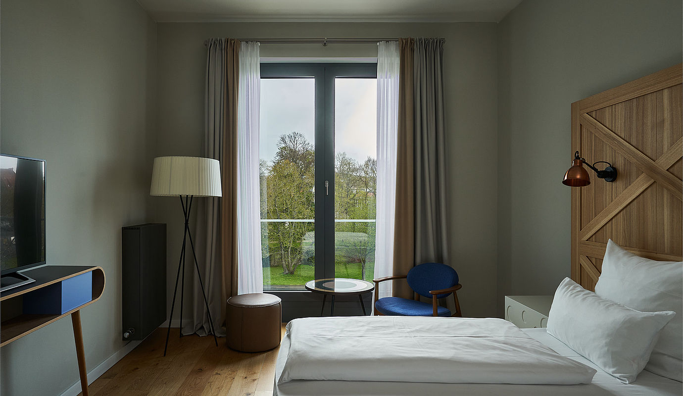 Blick auf ein großes Bett, einen Fernseher und ein helles Fenster in einem Hotelzimmer im FREIgeist