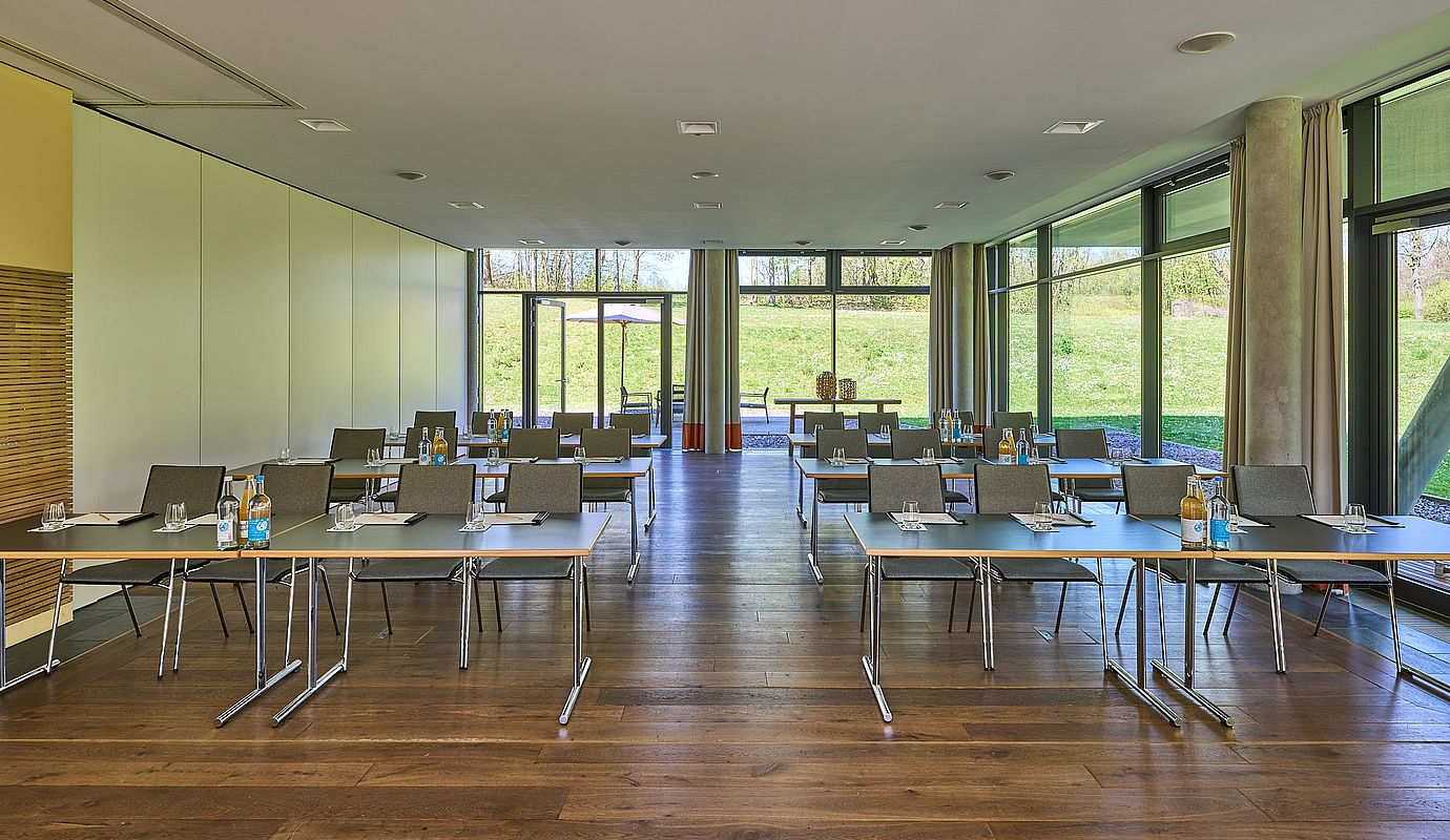 Zwei Reihen von Tischen und Stühlen in einem Konferenzraum mit großen Fenstern und Blick auf einen Garten