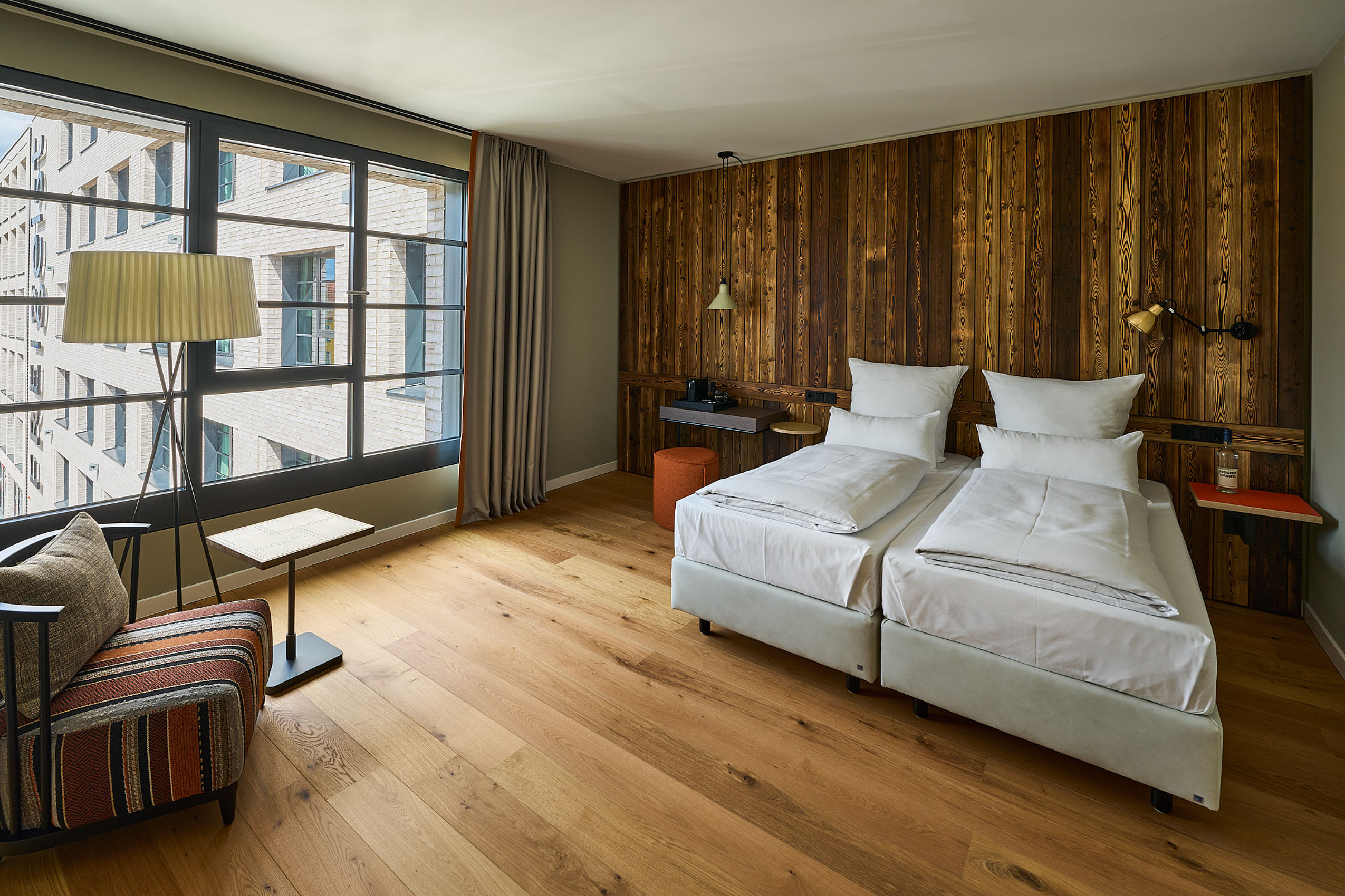 Ein schönes Hotelzimmer des FREIgeist in Göttingen Nordstadt mit einer dunklen Holzwand, einem frisch gemachten Bett, einer Stehlampe und großen Fenstern