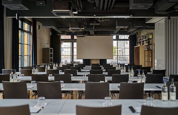 Stühle und Tische, die auf einen Wandprojektor in einem Konferenzraum des Hotels FREIgeist Göttingen Nordstadt gerichtet sind