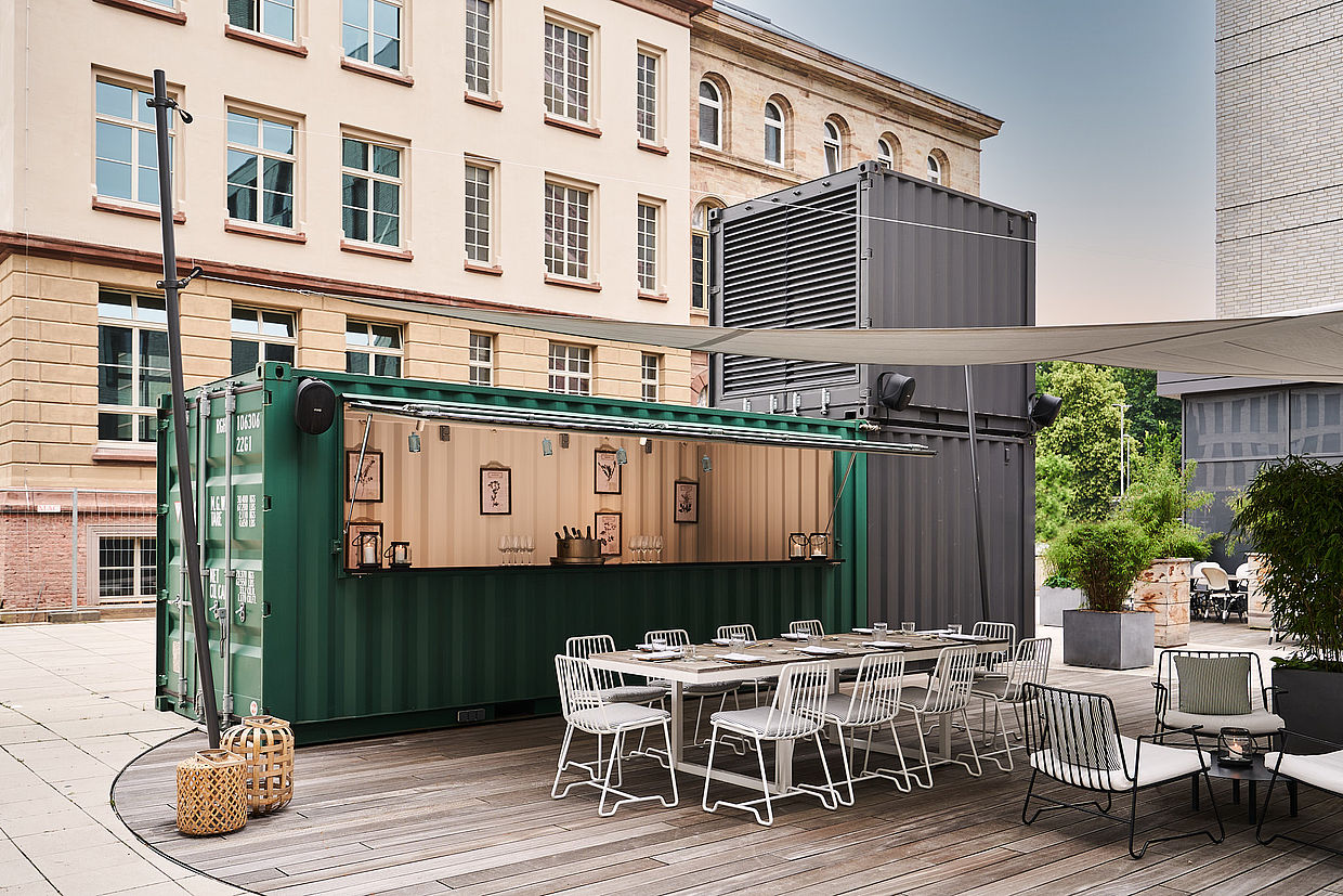 Eine Außenbar aus grünen Containern mit weißen Stühlen und einem Tisch daneben