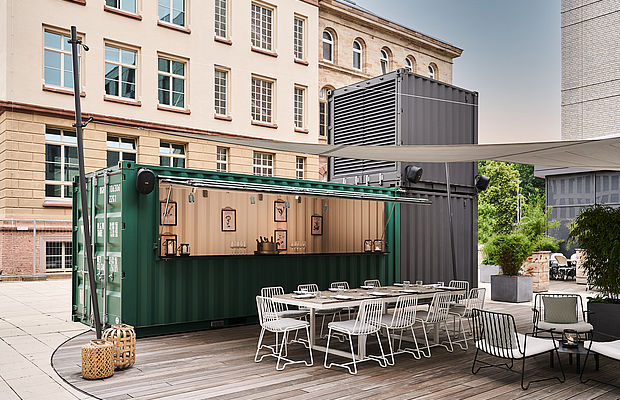 Eine Außenbar aus grünen Containern mit weißen Stühlen und einem Tisch daneben