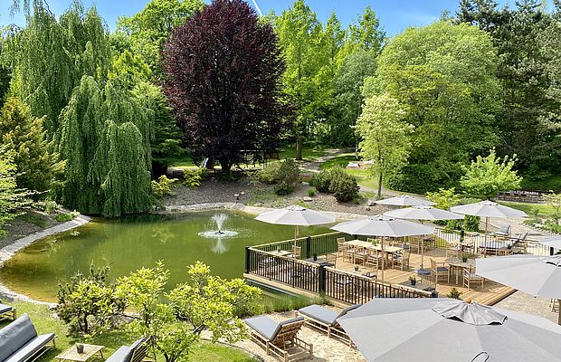 Holzterrasse mit Tischen, Stühlen und Sonnenschirmen über einem Teich.
