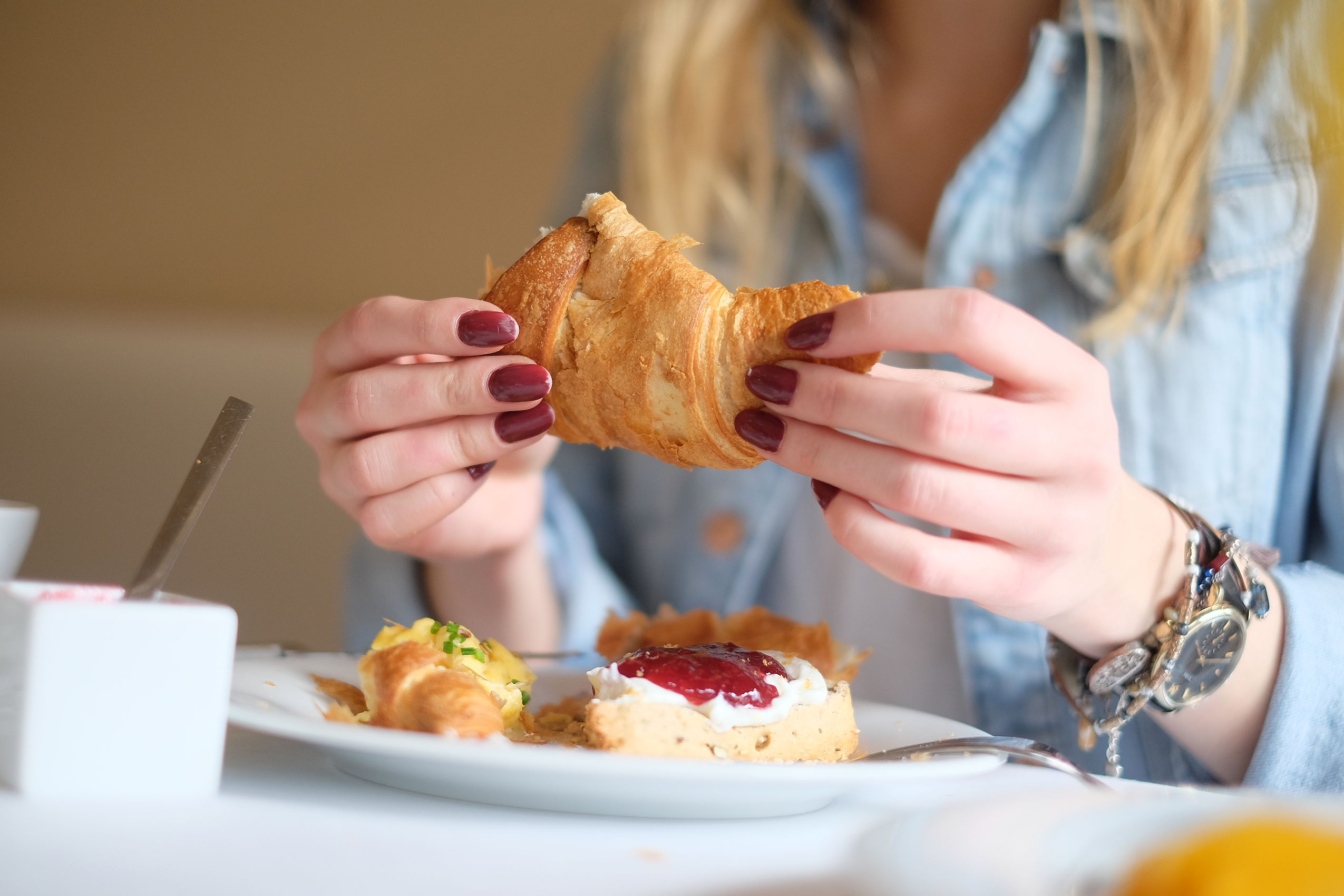Eine Frau mit roten Fingernägeln sitzt am Frühstückstisch und isst ein leckeres Croissant mit Marmelade