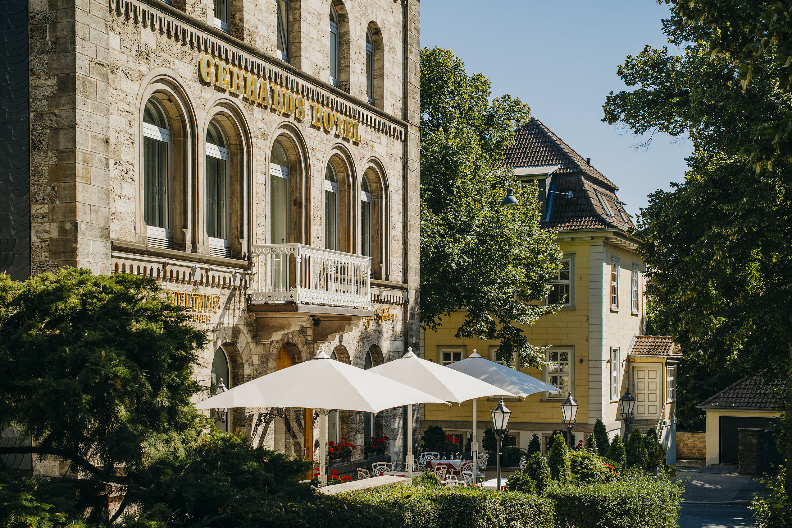 Sonnige Außenansicht des Gebhards Hotels und der Sonnenterasse mit geöffneten Schirmen und goldenem Hotelschriftzug an der Fassade