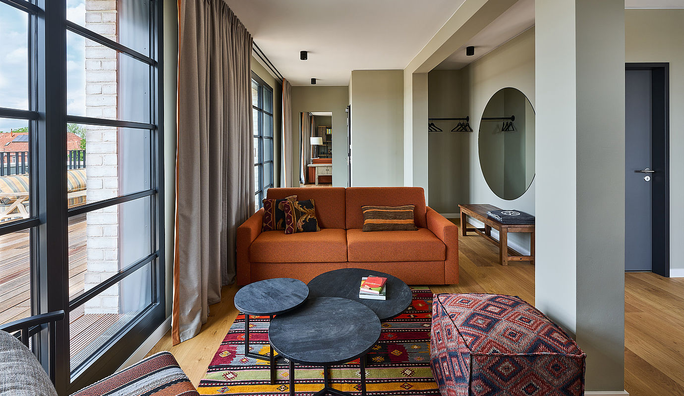 Ein Hotelzimmer mit orangener Couch und tiefen Fenstern mit Ausblick auf die Sonnenterrasse