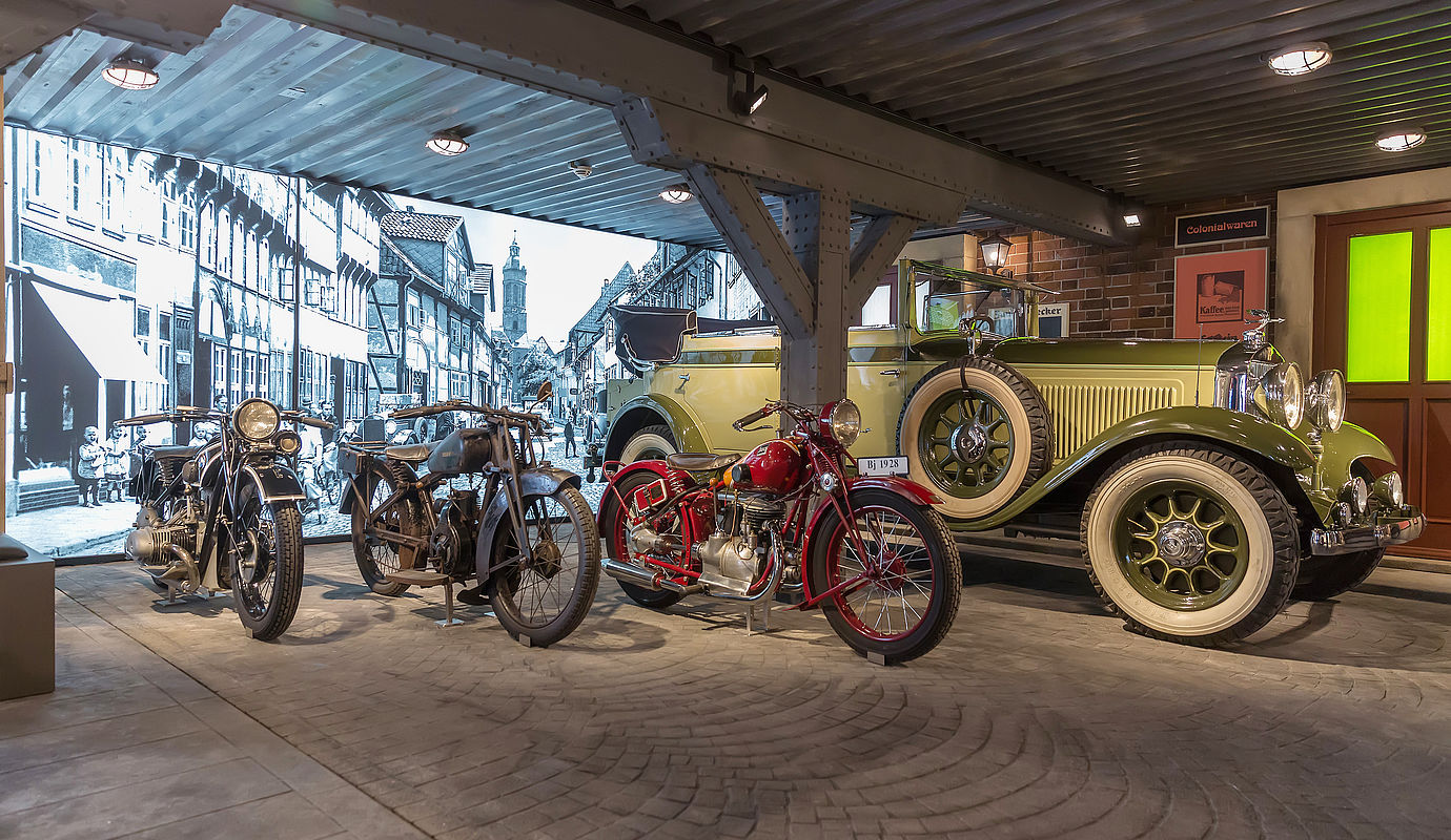 Drei Motorräder und ein Oldtimer ausgestellt vor einer schwarz-weißen Wand