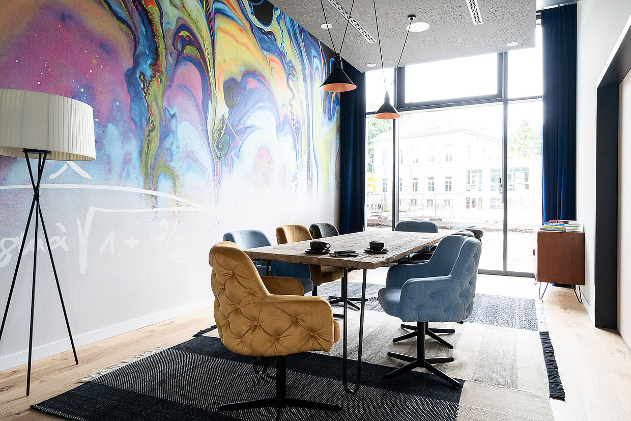 Ein langer Tisch mit bequemen Stühlen vor einer bunten Wand eines Meetingraums