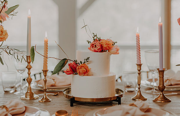 Eine Nahaufnahme einer Hochzeitstorte mit orangefarbenen Blumen, daneben Kerzen und Dekorationen