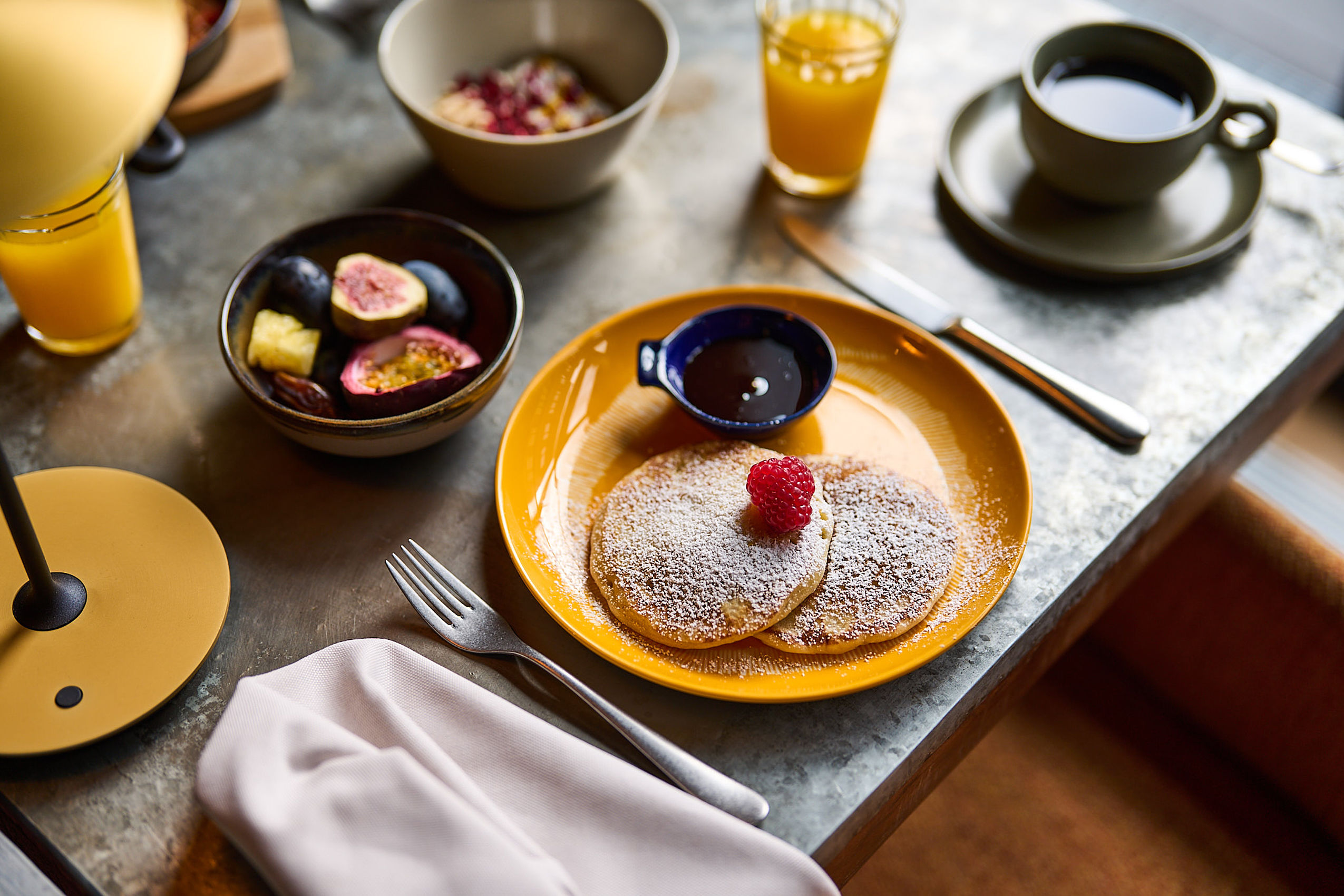 Zwei Pancakes mit Ahornsirup auf einem gelben Teller, dazu Feigen und eine Tasse Kaffee