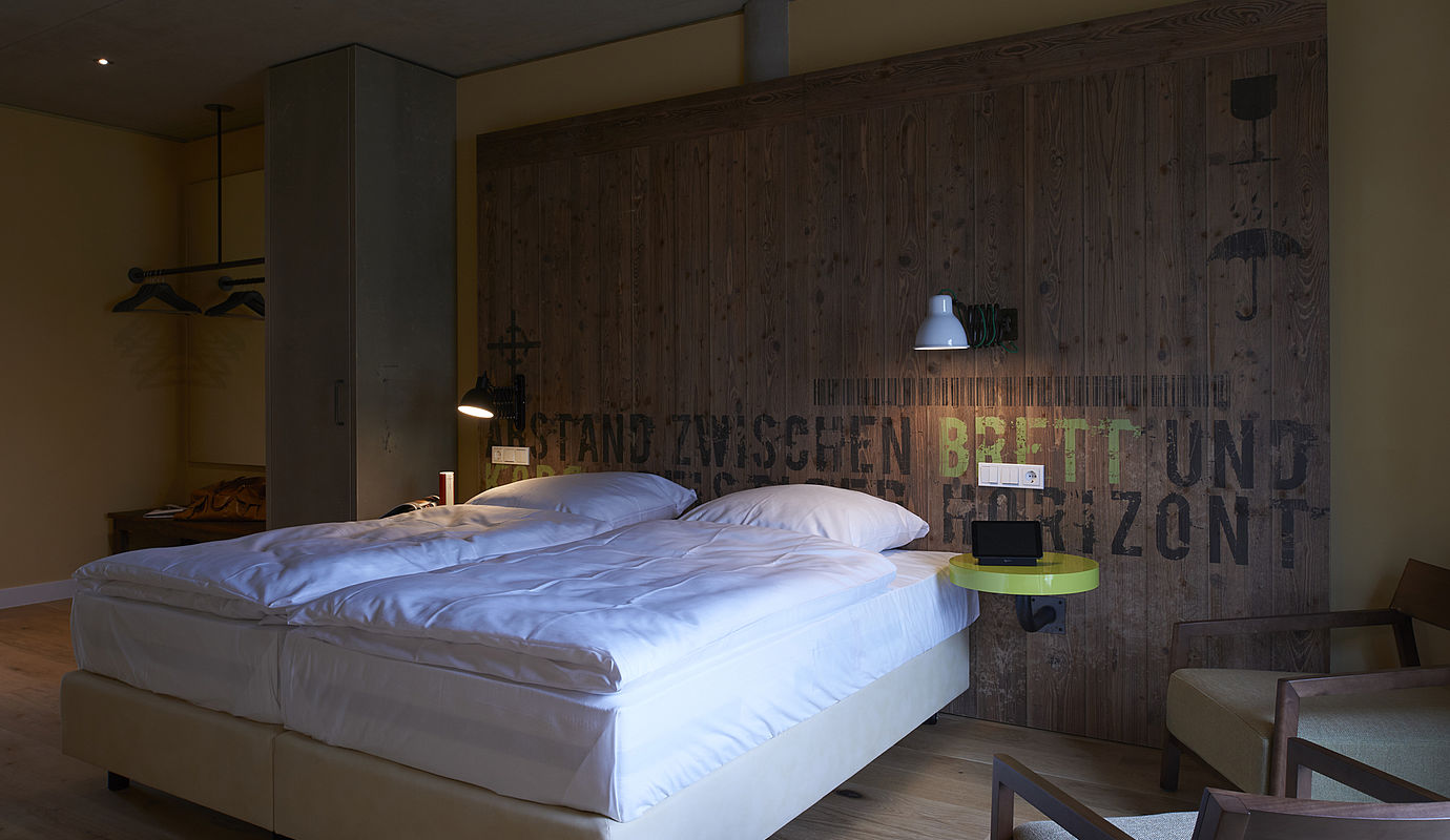 Ein großes Bett steht an einer bedruckten Holzwand an der zwei leuchtende Nachttischlampen hängen