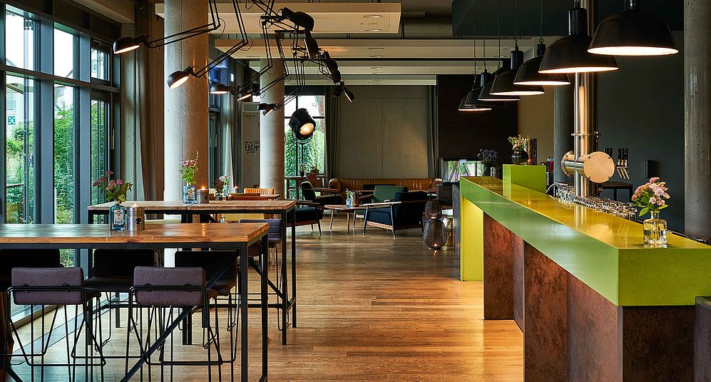 Blick auf die Hotelbar mit grünem Tresen und hohen Tischen und Hockern an einer Glasfront