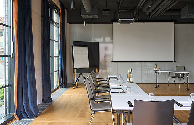 Konferenztische, Stühle und ein Projektor in einem hellen Konferenzraum mit großen Fenstern