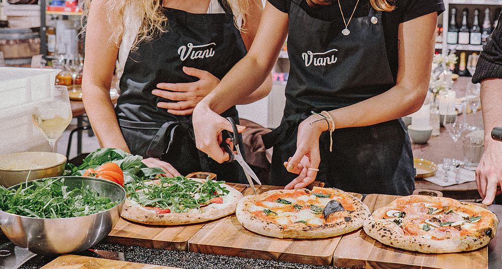 Zwei Personen in Viani-Schürzen schneiden eine Pizza an einem Tisch mit drei Pizzen.