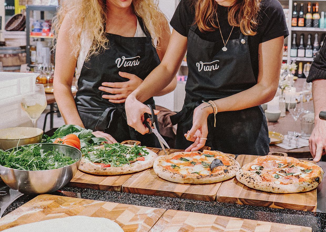 Zwei Personen in Viani-Schürzen schneiden eine Pizza an einem Tisch mit drei Pizzen.